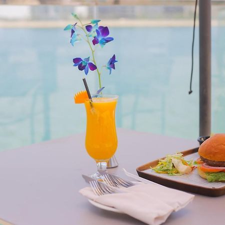 Grand Swiss-Belhotel Waterfront Seef Sanabis Exterior foto The photo features a refreshing scene with a vibrant orange drink garnished with a slice of orange and purple flowers, served in a glass. Next to it, there is a tray containing a hamburger with lettuce and possibly other condiments, accompanied by a 