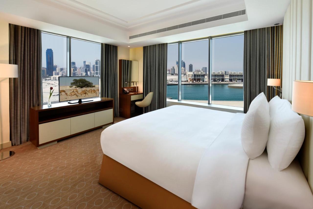 Grand Swiss-Belhotel Waterfront Seef Sanabis Exterior foto The photo shows a luxurious hotel room featuring a large bed with white pillows and a neatly made bedspread. The room has floor-to-ceiling windows that offer a view of a city skyline, suggesting a vibrant urban setting. There is a modern television m