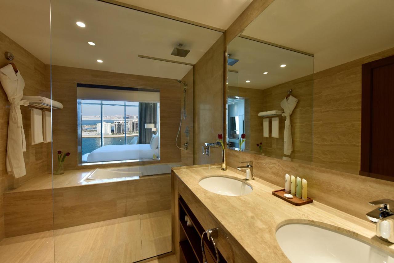 Grand Swiss-Belhotel Waterfront Seef Sanabis Exterior foto The photo depicts a modern bathroom with a sleek design. It features a large mirror above a double sink with a wooden cabinet below. The walls are adorned with light-colored tiles, and there is a glass shower enclosure. On one side of the room, there