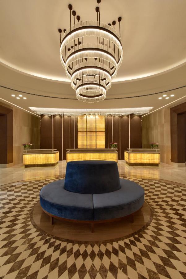 Grand Swiss-Belhotel Waterfront Seef Sanabis Exterior foto The photo shows an elegant hotel lobby. It features a circular seating area in the center with a plush, dark blue upholstered bench. Surrounding this seating area is a patterned floor made of light and dark tiles. Behind the seating area, there are t