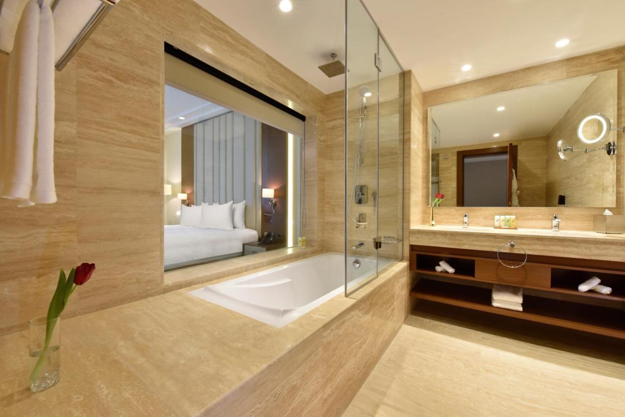 Grand Swiss-Belhotel Waterfront Seef Sanabis Exterior foto The photo shows a modern bathroom with a sleek design. It features a bathtub, which is partially enclosed by glass. The walls are made of light-colored stone, giving a clean and elegant look. There is a large mirror above the vanity, which has some l