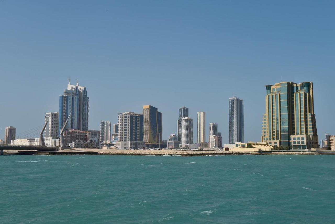 Grand Swiss-Belhotel Waterfront Seef Sanabis Exterior foto The photo shows a skyline consisting of several tall buildings and skyscrapers along a waterfront. The buildings vary in design and height, showcasing modern architecture typical of urban areas. The water in the foreground is a vibrant turquoise colo