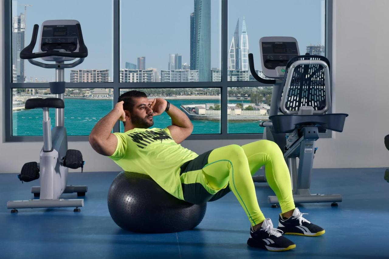 Grand Swiss-Belhotel Waterfront Seef Sanabis Exterior foto The photo shows a man exercising in a gym setting. He is performing a sit-up while balancing on an exercise ball. He is wearing a bright neon yellow athletic outfit and black shoes. In the background, large windows provide a view of a city skyline an