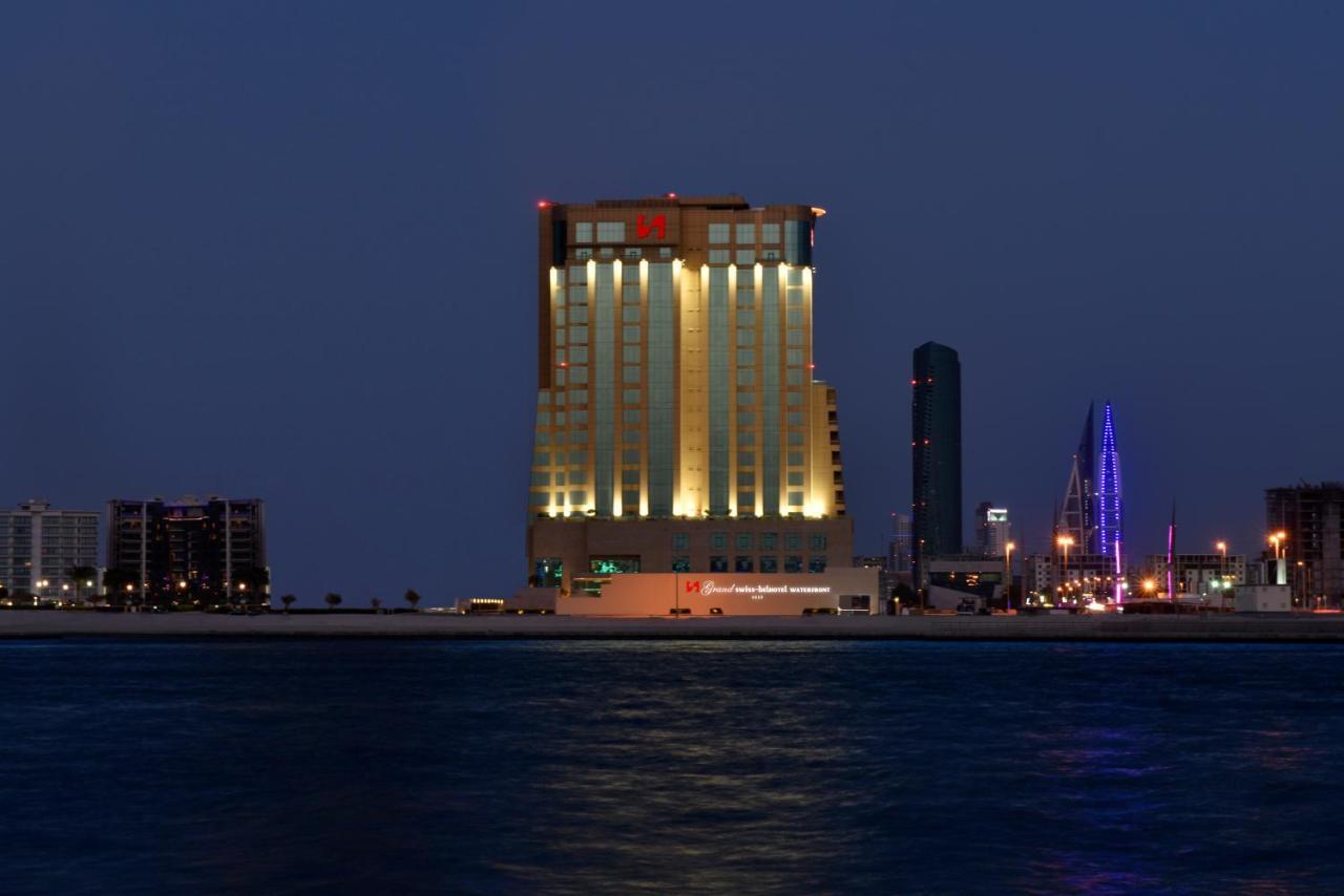 Grand Swiss-Belhotel Waterfront Seef Sanabis Exterior foto The photo shows a tall building illuminated at night, with a blend of warm yellow and cool white lights. The structure has a modern architectural design and appears to be a hotel or high-rise establishment. It is situated near a body of water, likely