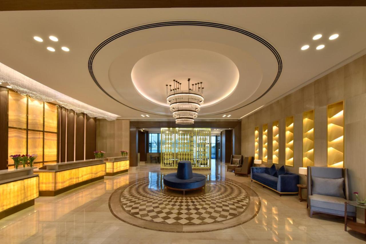 Grand Swiss-Belhotel Waterfront Seef Sanabis Exterior foto The image depicts a modern hotel lobby. It features a stylish design with warm lighting and elegant decor. There is a reception desk made of light-colored wood and illuminated panels. The ceiling has a large, circular light fixture, adding a luxuriou