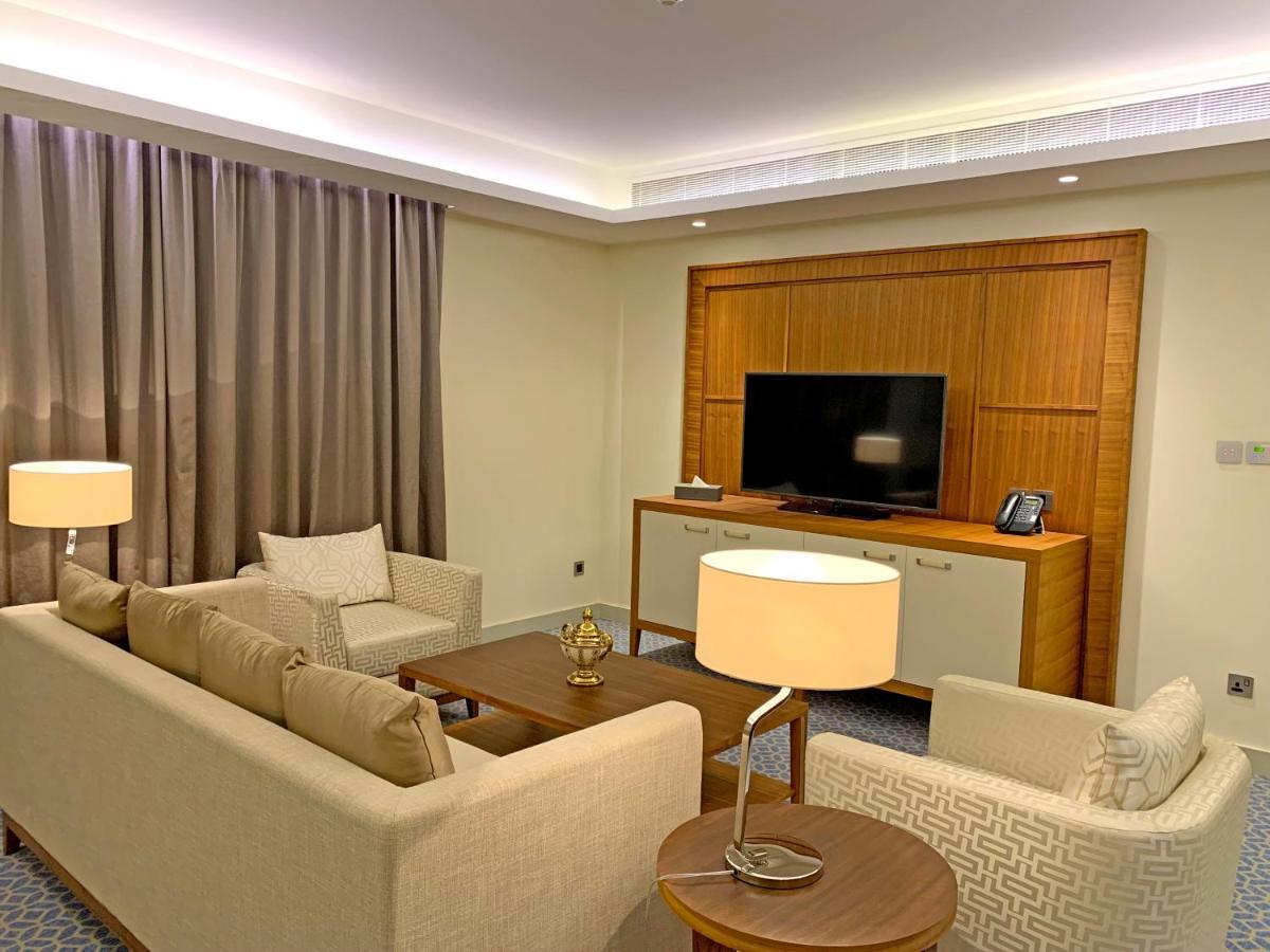 Grand Swiss-Belhotel Waterfront Seef Sanabis Exterior foto The photo shows a modern living room area. There are two sofas arranged around a coffee table, which has a decorative item on it. A lamp is positioned next to one of the sofas, providing ambient lighting. In the background, there is a large flat-scre