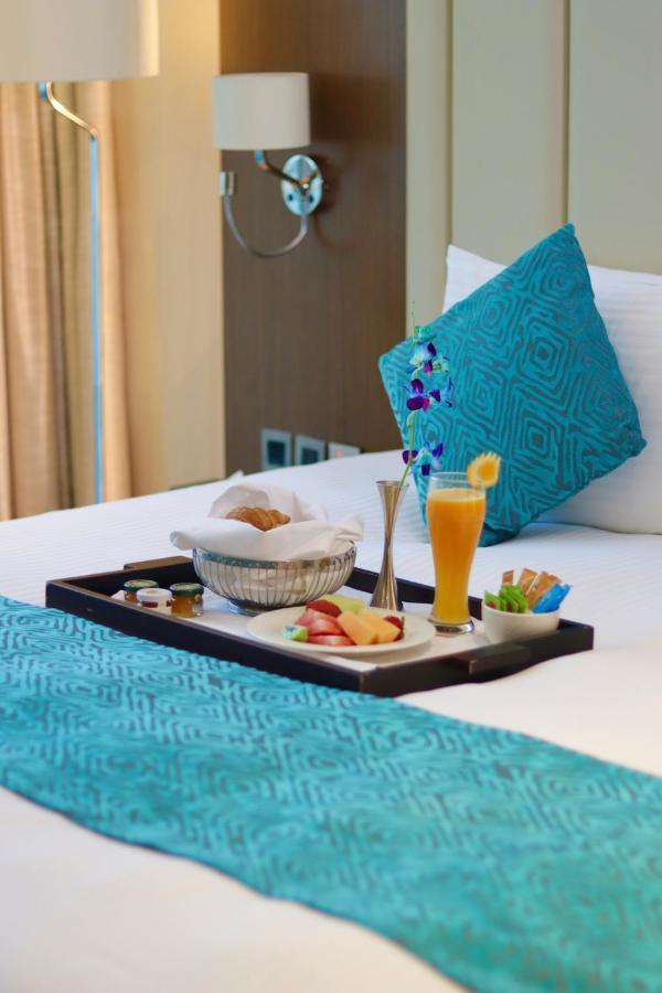 Grand Swiss-Belhotel Waterfront Seef Sanabis Exterior foto The photo shows a tray placed on a bed. On the tray, there is a variety of food items, including a basket with a pastry, sliced fruits, and small jars, likely containing condiments or spreads. There is also a tall glass filled with a bright orange be