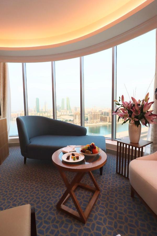 Grand Swiss-Belhotel Waterfront Seef Sanabis Exterior foto The image shows a stylish and modern interior of a hotel room or lounge area. There is a curved blue sofa positioned near large windows that offer a view of a cityscape, featuring tall buildings and what appears to be water in the foreground. A round