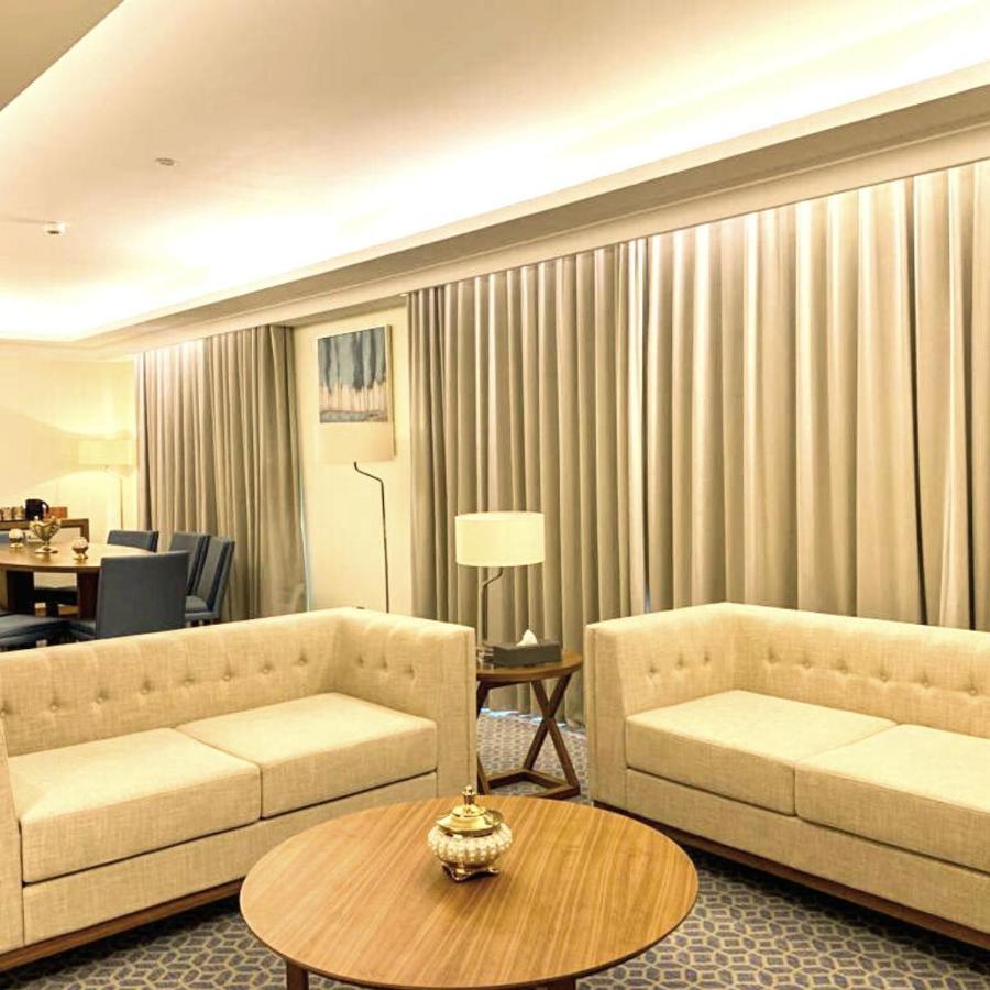 Grand Swiss-Belhotel Waterfront Seef Sanabis Exterior foto The photo shows a modern, softly lit living area featuring two light-colored sofas with simple, elegant designs. In the center, there is a round wooden coffee table adorned with a decorative item. The walls are adorned with large, neutral-toned curta