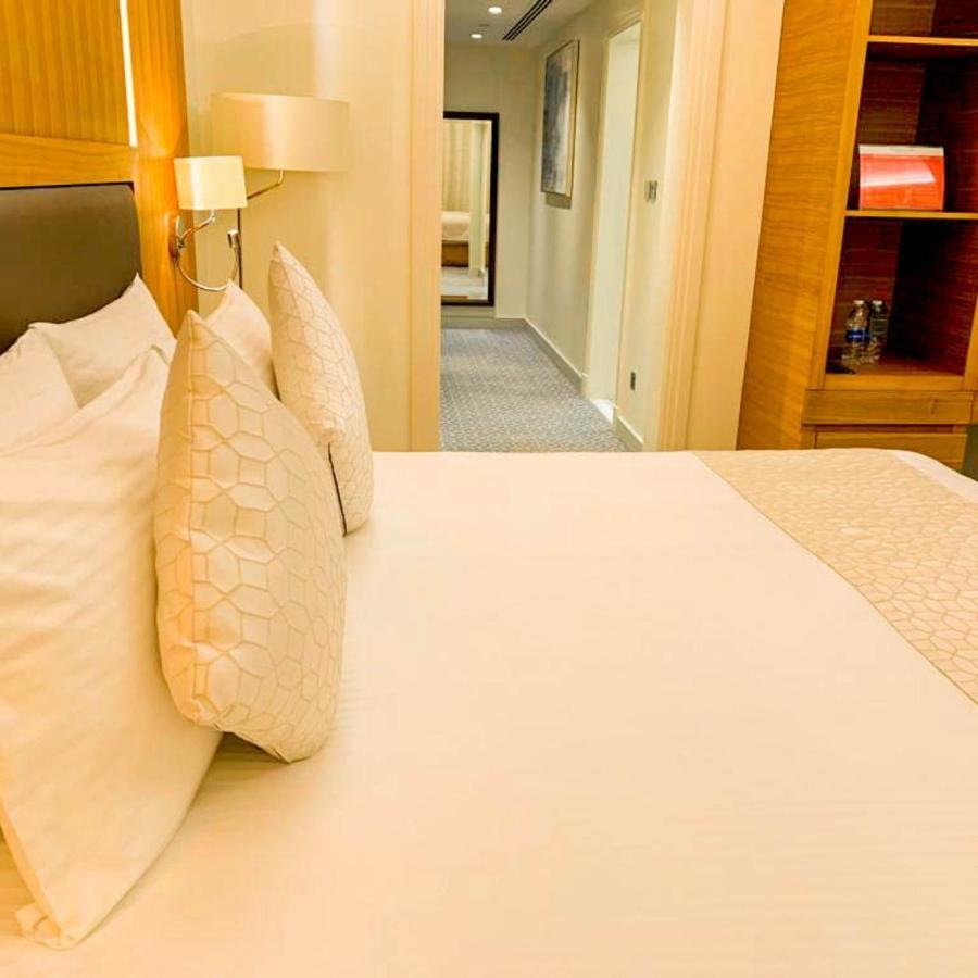 Grand Swiss-Belhotel Waterfront Seef Sanabis Exterior foto The photo shows a neatly made bed with a light-colored blanket and decorative pillows. In the background, there is a hallway leading to other room areas. The walls are painted in neutral tones, and there are elements of modern decor, such as a bedsid