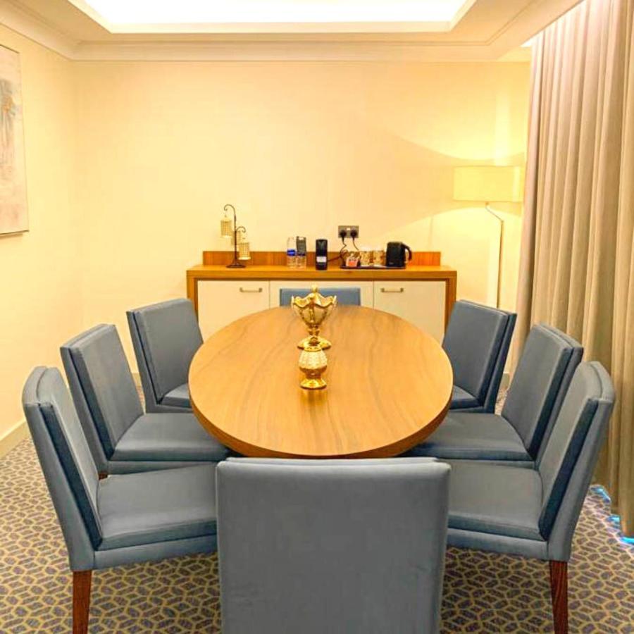 Grand Swiss-Belhotel Waterfront Seef Sanabis Exterior foto The photo shows a modern dining or conference room setup. It features a round wooden table surrounded by several blue upholstered chairs. In the center of the table, there is a decorative golden piece, likely a vase or trophy. To one side of the room