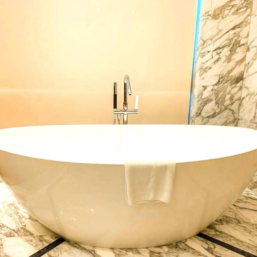 Grand Swiss-Belhotel Waterfront Seef Sanabis Exterior foto The photo shows a modern, oval-shaped bathtub located in a bathroom. The tub is white and has a sleek, minimalist design. A chrome faucet with a handle is mounted above the tub. There’s a white towel draped over the edge of the tub. The surrounding w