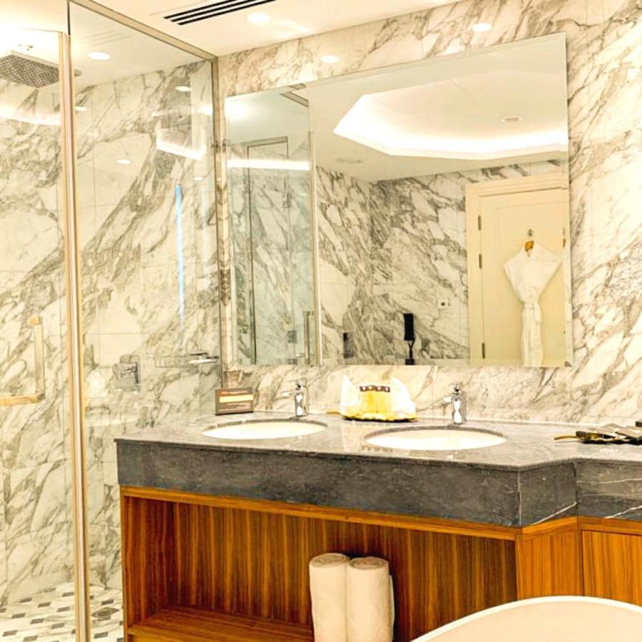 Grand Swiss-Belhotel Waterfront Seef Sanabis Exterior foto The photo depicts a luxurious bathroom featuring elegant marble walls. There are two sinks with a dark granite countertop, and the cabinetry below has a warm wooden finish. Large mirrors are mounted above the sinks, reflecting the stylish design. A w